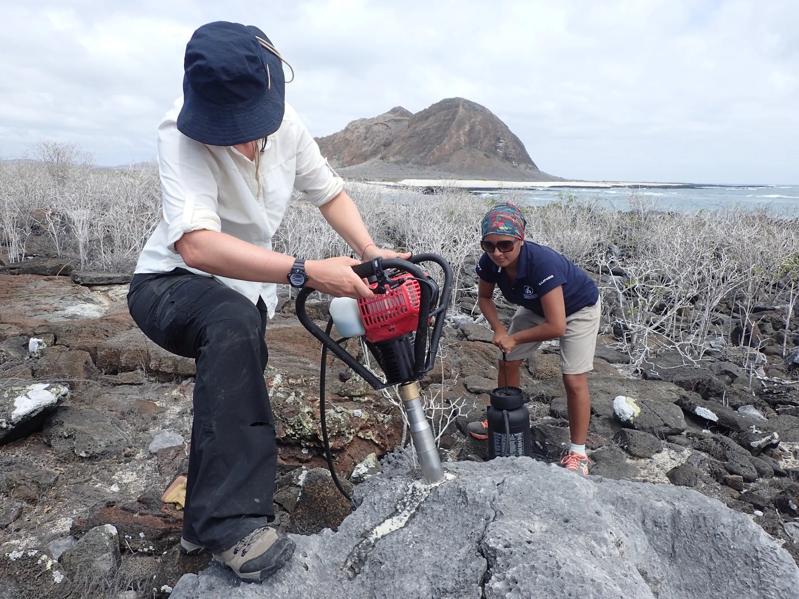 One person stands with a pneumatic drill that is drilling into a gray fossilized coral boulder. Their foot is braced on the boulder and their face is blocked by a hat. Another scientist stands crouched behind watching.