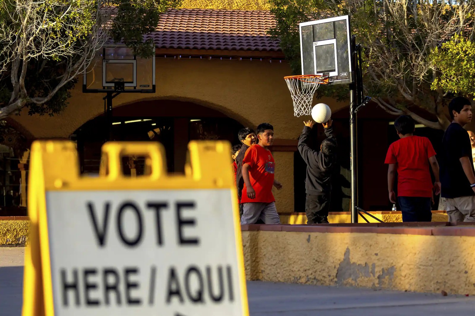 En primer plano se ve un letrero que dice «Vote aquí / Aquí», mientras unos jóvenes juegan baloncesto frente a un edificio al fondo.