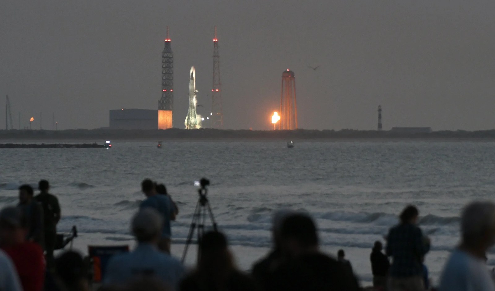 Shadowy spectators in the foreground watch a large rocket on a launch pad in the distance under a dark gray sky.