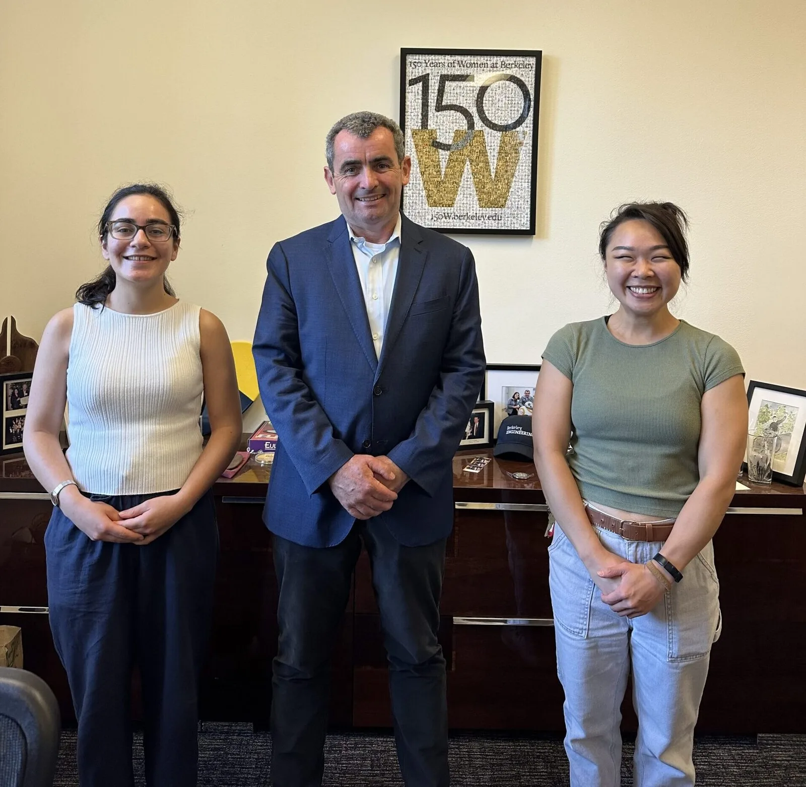 A group photograph of three smiling people standing side-by-side in an office setting. On the left is a woman in a white ribbed sleeveless top and dark trousers. In the center stands a man in a blue blazer over a white shirt, with clasped hands. On the right is a woman in an olive green t-shirt and light grey cargo pants. In the background is a wooden credenza covered in framed pictures, and above it, a framed poster on the wall that reads "150 W"