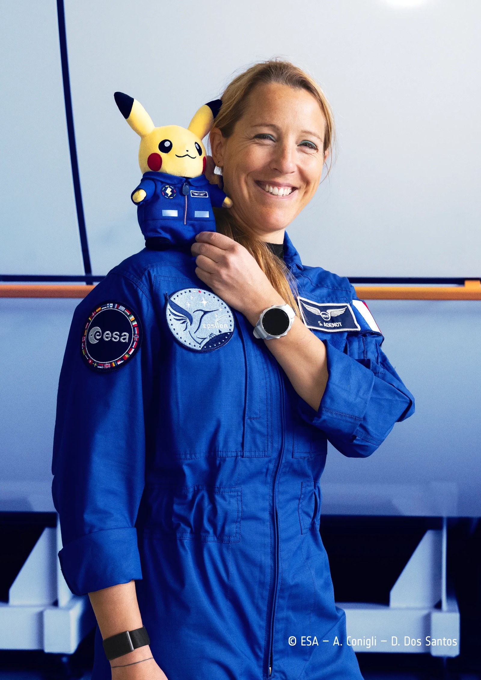 an ESA astronaut in a blue flight suits smiles with a plush pikachu on her shoulder