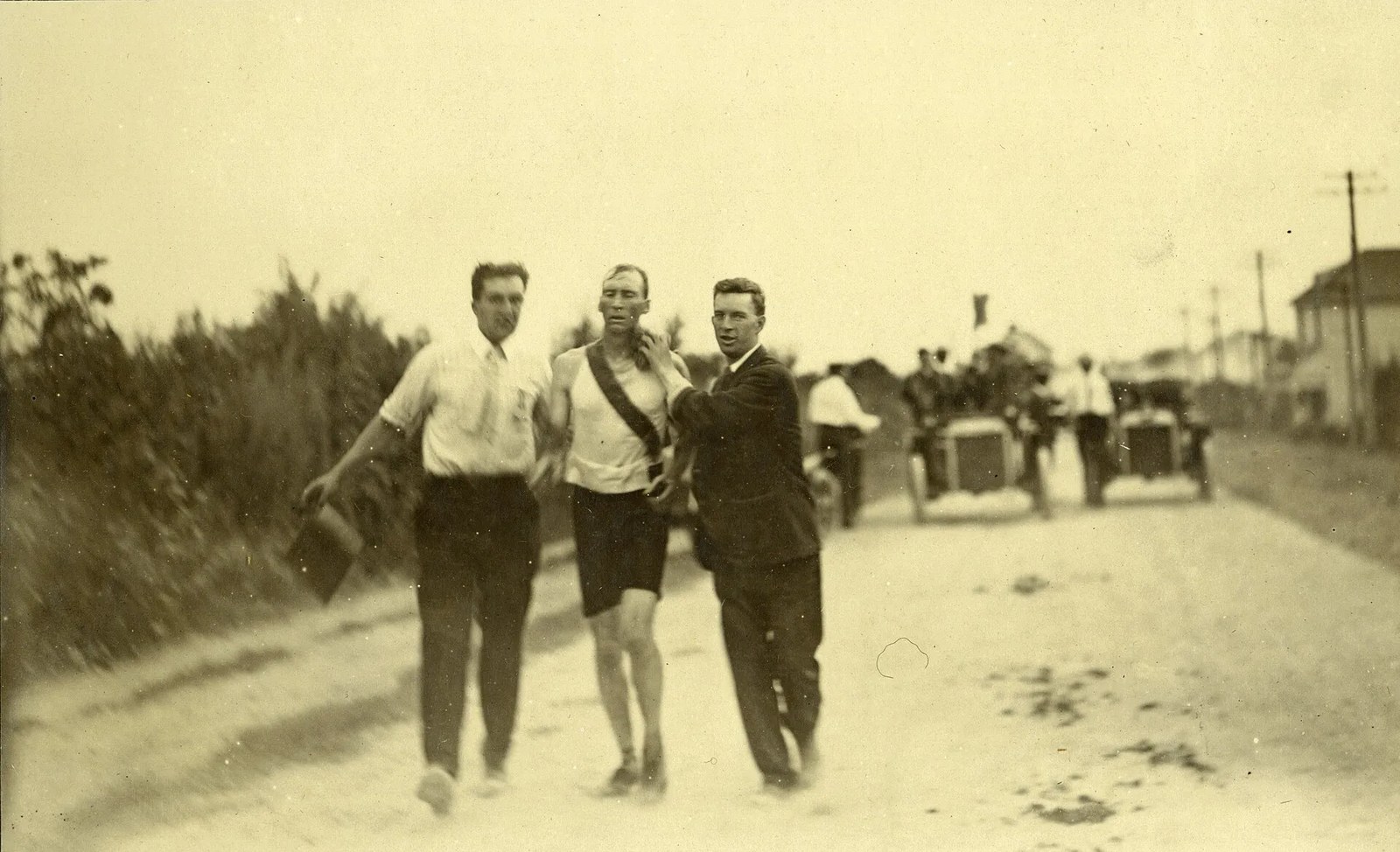 Historic photo showing two men next to a runner try to help him with vintage cars in the background.
