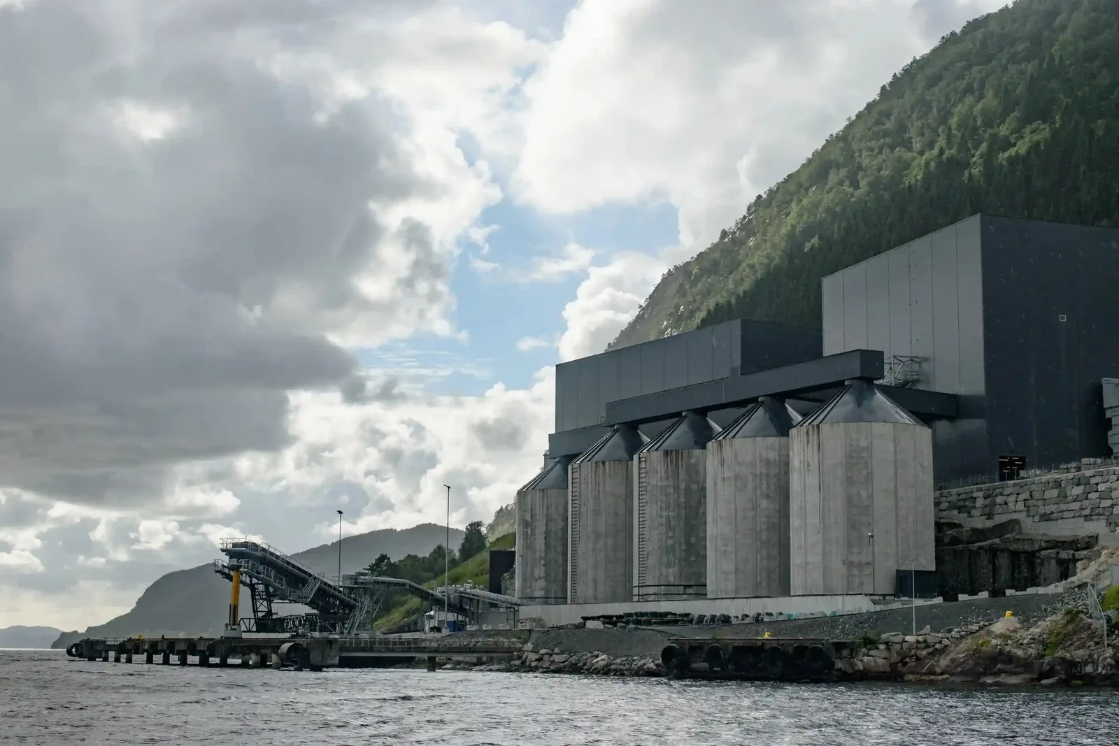 A mining plant beside a body of water against a steep tree-covered mountainside