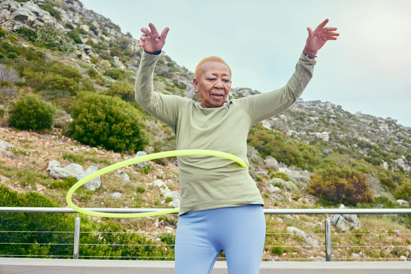 A dynamic, medium-low angle shot of an elderly woman with short, orange-blonde hair, actively hula hooping outdoors on a wooden deck. She wears a sage green long-sleeved shirt and light blue leggings. Her arms are raised and outstretched for balance, hands open, with a focused expression as a lime green hoop spins around her waist. She stands before a silver railing. The background features a vast, scrub-covered hillside with bushes and rocks under a bright, hazy sky.