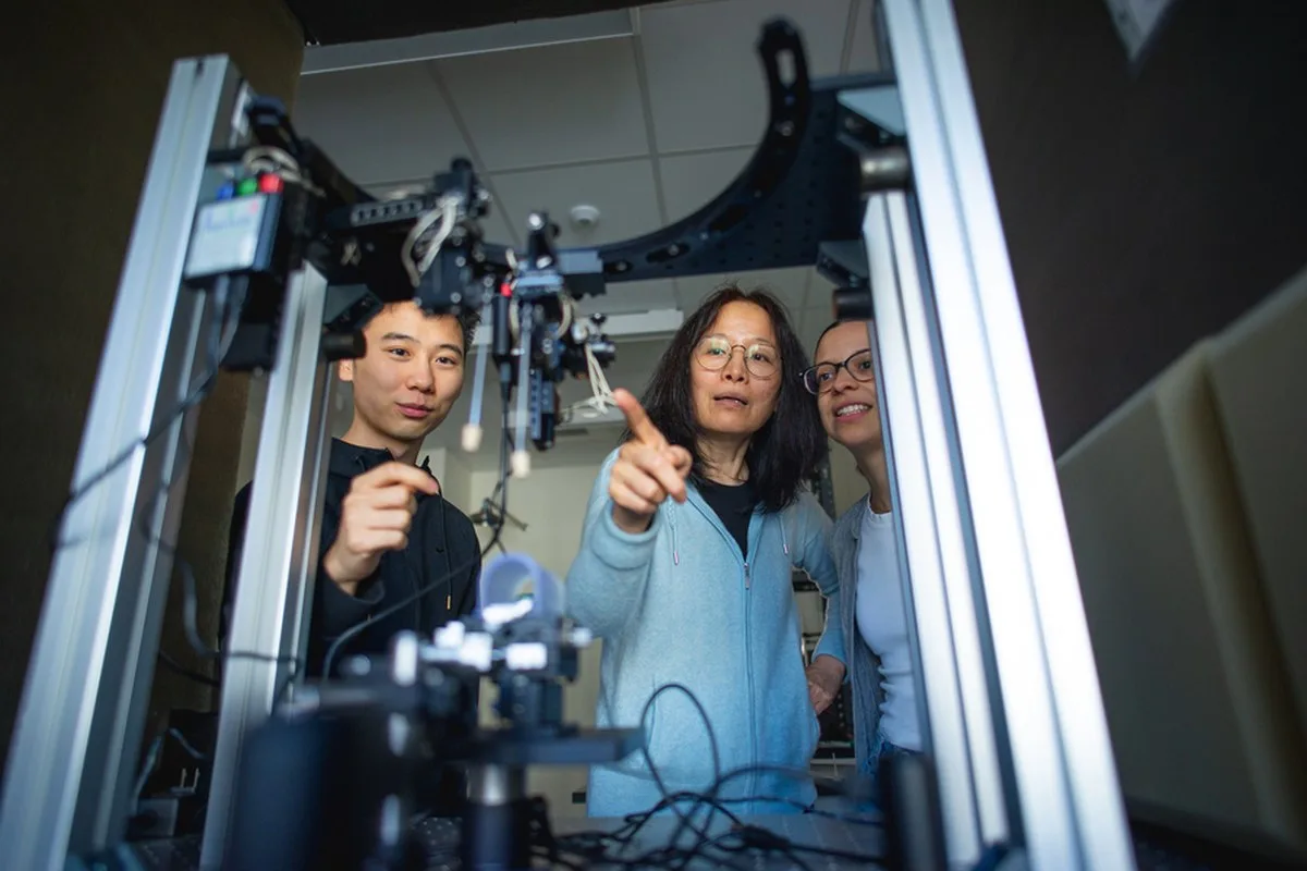 Fan Wang stands between two students, pointing to some lab hardware