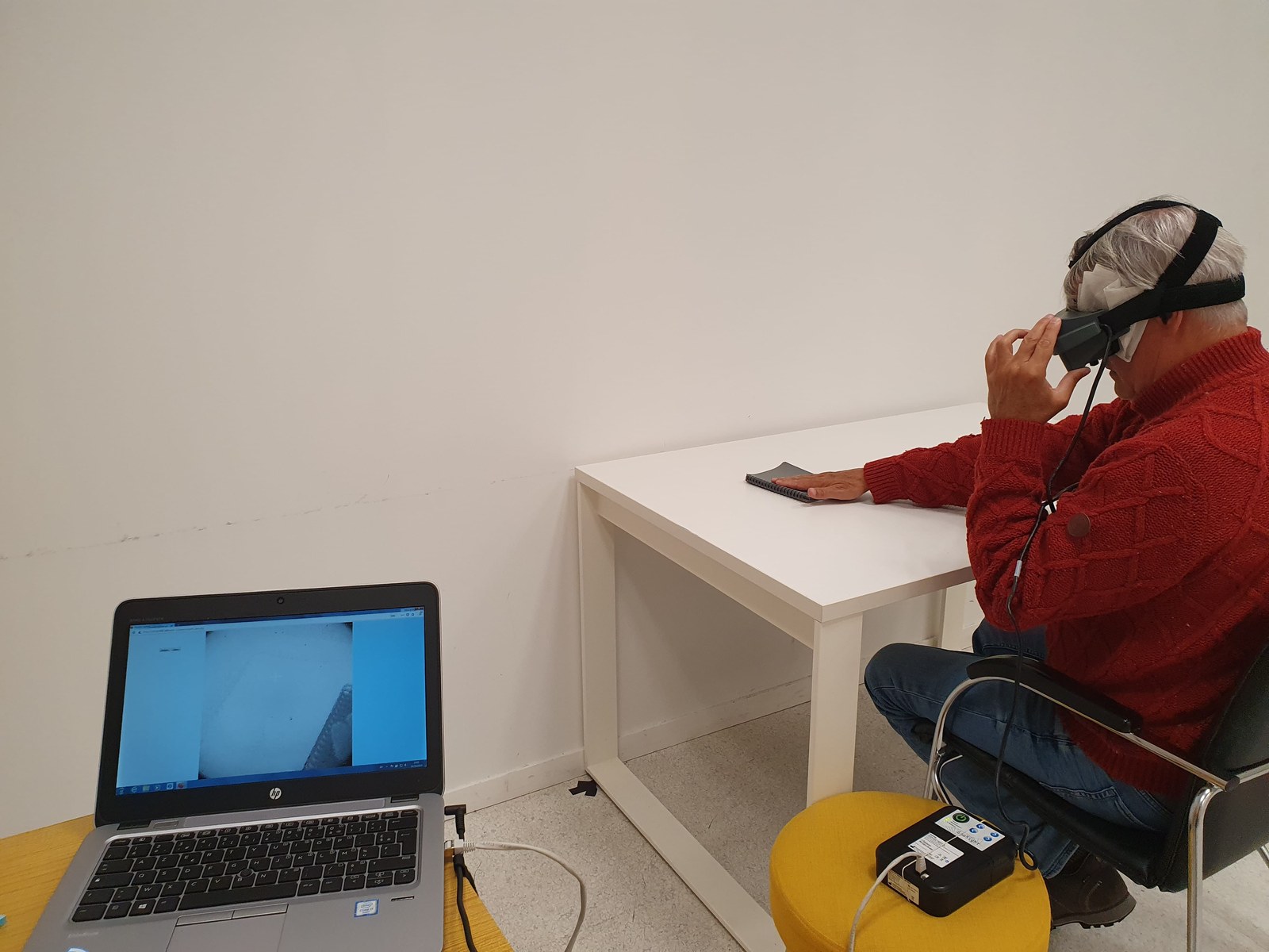 A seated participant wearing special goggles touches a notebook on a white table, with a laptop and connected equipment nearby.